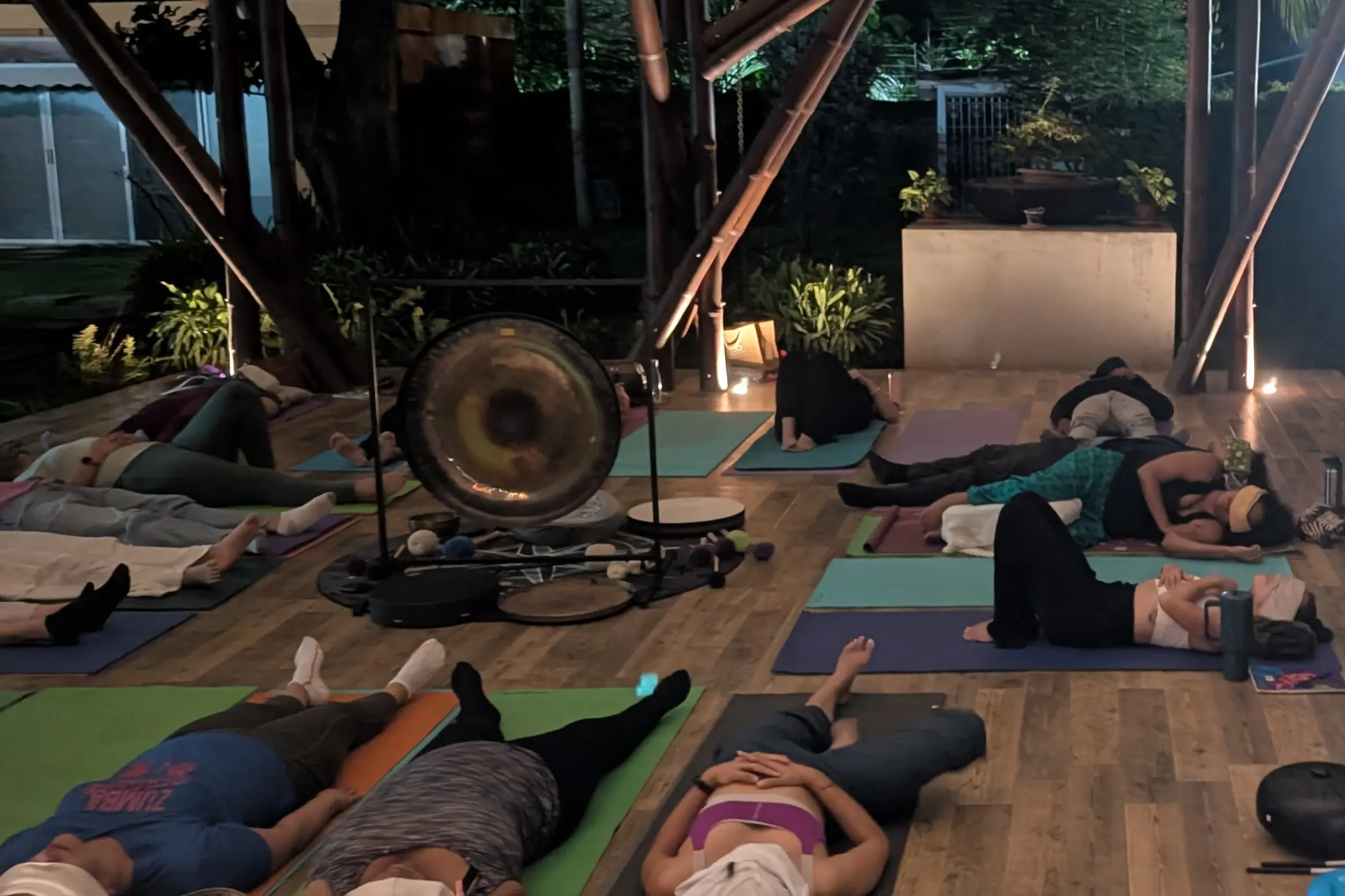 Un grupo de personas recostadas en colchonetas de yoga en un pabellón al aire libre por la noche, participando en una sesión de relajación o baño de sonido de aporte voluntario. En el centro hay un gran gong, cuencos tibetanos y otros instrumentos de percusión, rodeados de velas. La estructura tiene vigas de bambú y está iluminada con luz suave. La gente está cubierta con mantas y algunos llevan antifaces para dormir.