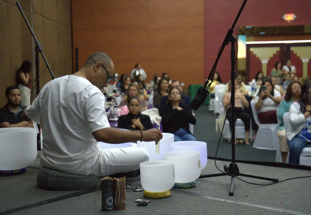 Un hombre vestido de blanco se sienta en un cojín en un escenario y toca una serie de cuencos de cristal blanco con un mazo durante una sesión de terapia de sonido. Hay micrófonos cerca de los cuencos para captar el sonido. Detrás de él, un grupo de personas sentadas en sillas escucha con atención, algunos con las manos sobre el pecho en señal de meditación, en un gran salón de eventos con iluminación tenue y paredes de madera.