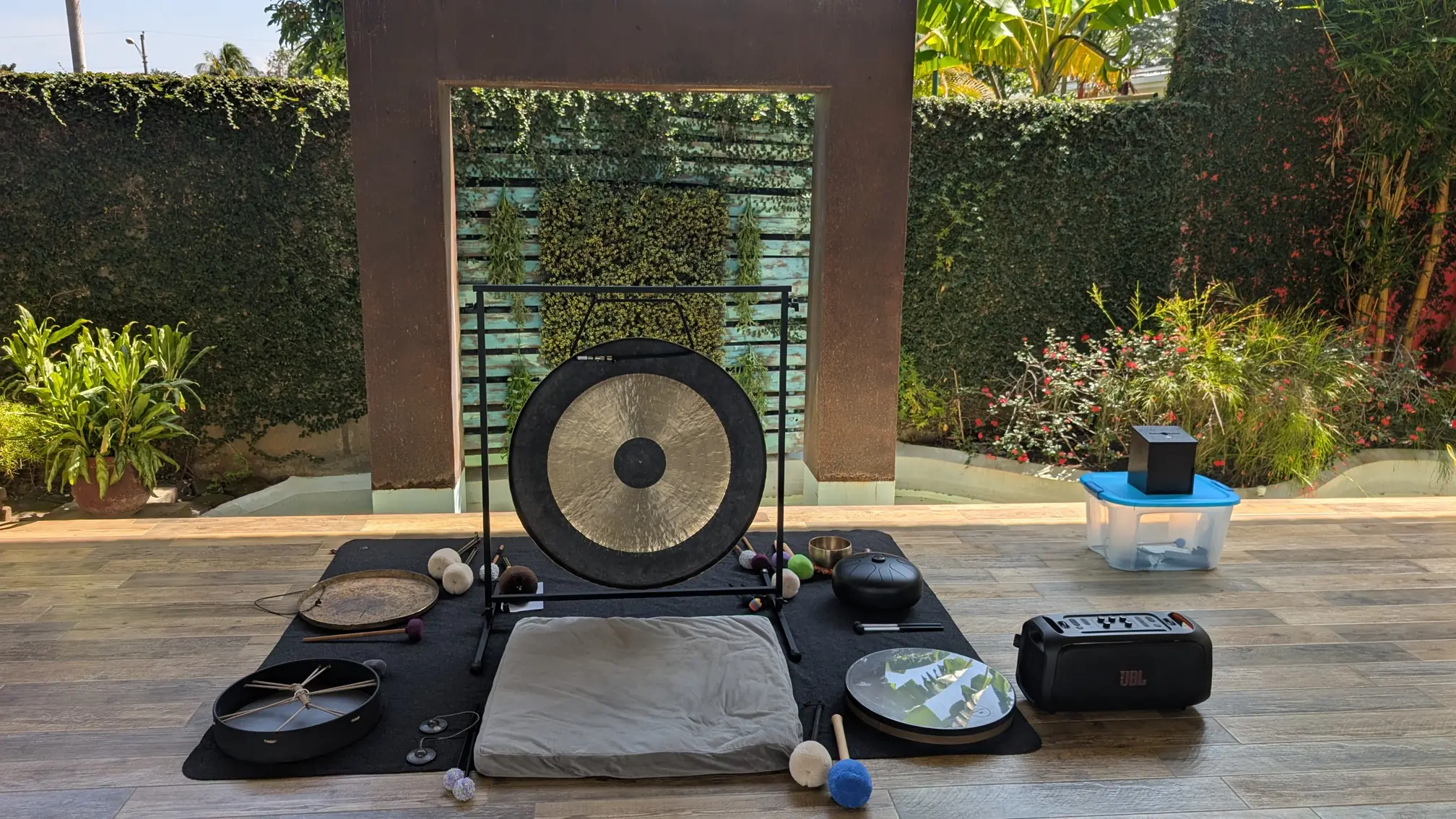 Una colección de instrumentos de sanación sonora dispuestos sobre una esterilla negra y un cojín gris en un patio soleado. En el centro destaca un gran gong en un soporte metálico. Alrededor hay un tambor chamánico, cuencos tibetanos, un tambor de lengüeta metálica (handpan), platillos tingsha, y varios mazos de fieltro de diferentes colores y tamaños. A la derecha, un altavoz portátil JBL y una caja de almacenamiento azul con una caja negra encima. El entorno incluye una pared de metal oxidado y una pared cubierta de hiedra, con un jardín exuberante al fondo.