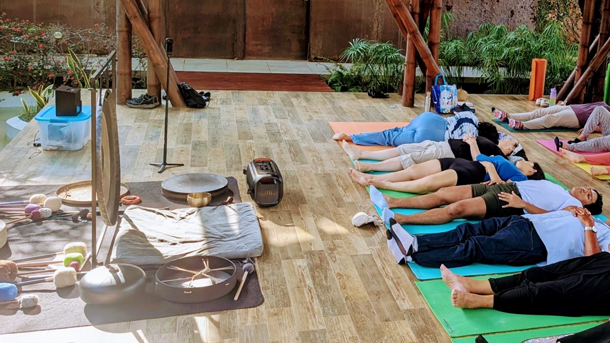 Esta imagen muestra un grupo de personas participando en una sesión de baño de sonido y meditación al aire libre en una terraza de madera cubierta
